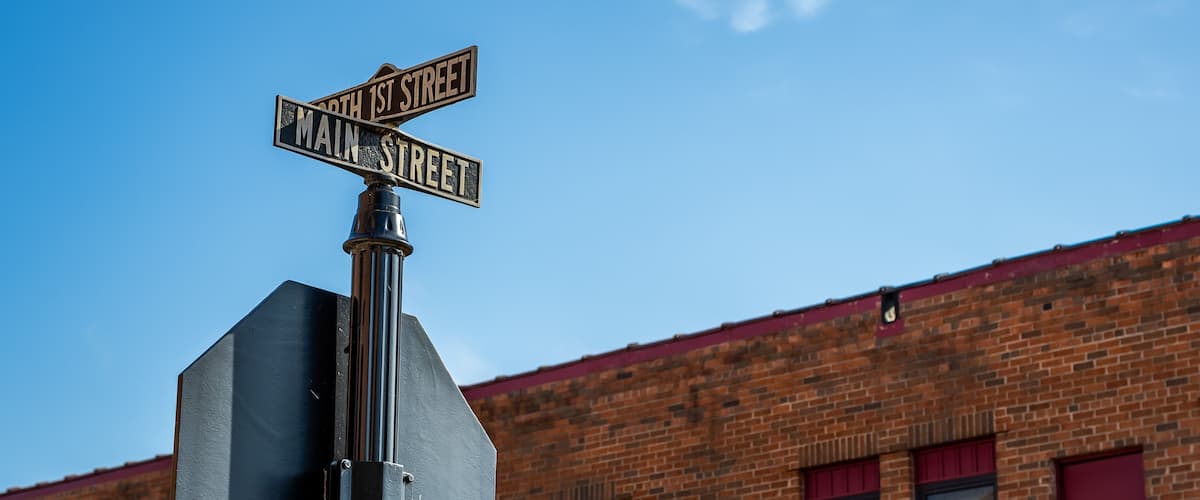 A street sign with 'North 1st Street' and 'Main Street' written on it, mounted on a metal pole, with a clear blue sky and a brick building in the background.