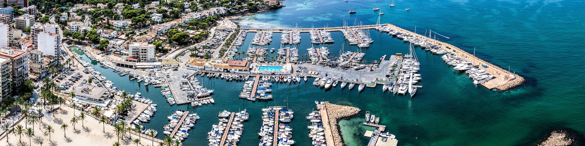 An aerial view of a coastal marina with numerous boats docked, a sandy beach, and a backdrop of residential buildings.