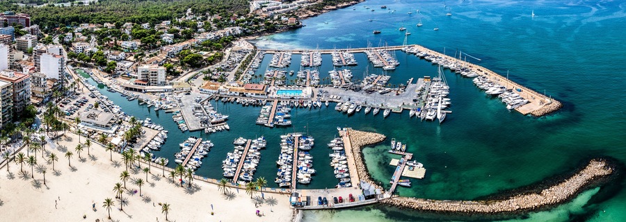 Vista aérea de un puerto costero con numerosas embarcaciones amarradas, una playa de arena y edificios residenciales al fondo.