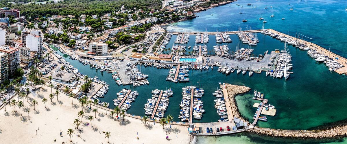 An aerial view of a coastal marina with numerous boats docked, a sandy beach, and a backdrop of residential buildings.