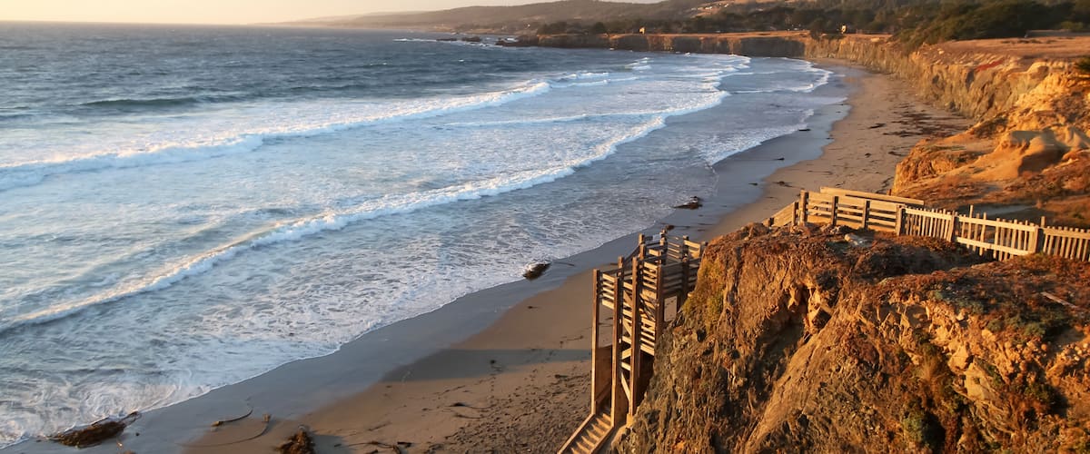 A coastal beach with waves, a wooden staircase, and a rocky cliff.