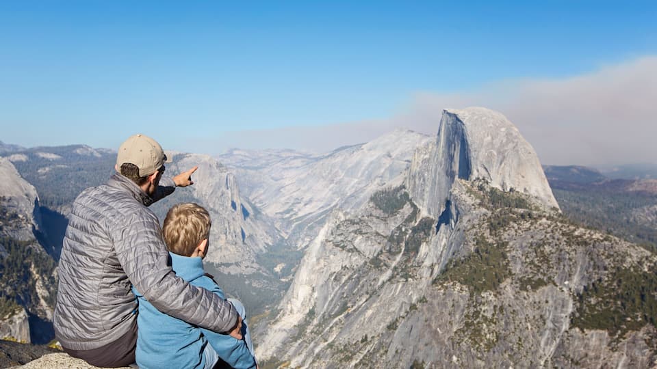 A man and a child sitting on a rocky ledge overlooking a mountainous landscape with a prominent rock formation.