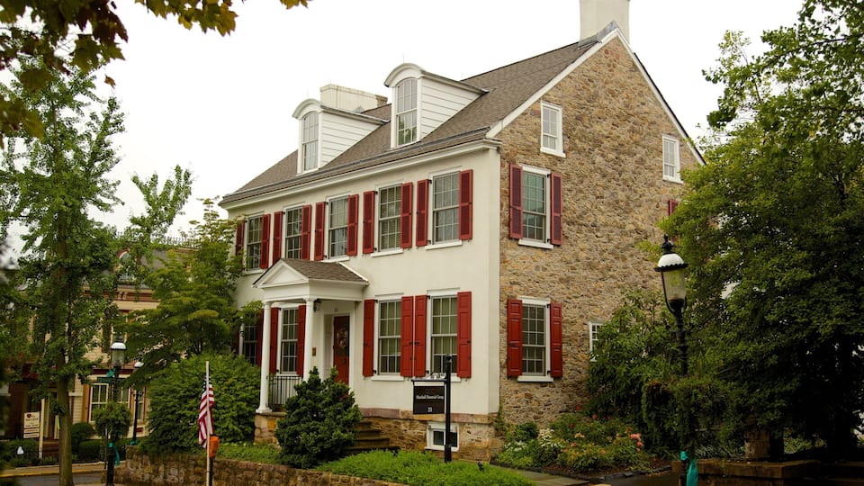 A historic stone building with red shutters and a white facade, surrounded by lush greenery.