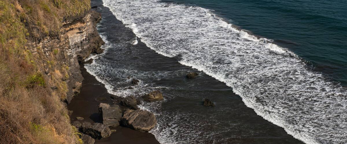 Aerial view of white foamy ocean waves, black volcanic sandy beaches and rocky cliffs. Sunny day, blue sky. Pacific coast of El Salvador.