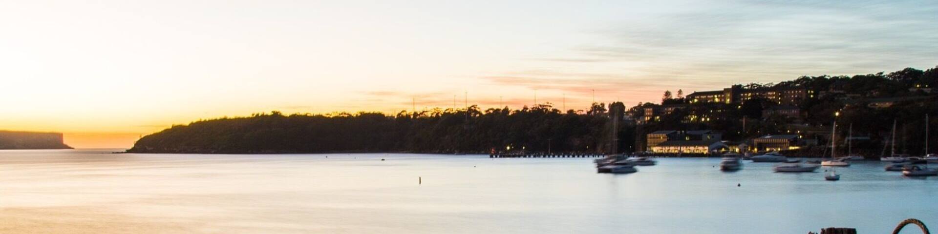 A rocky shoreline with a rope tied to a post, calm water, and boats in the distance during sunset.