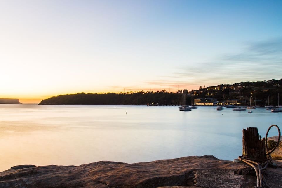 A rocky shoreline with a rope tied to a post, calm water, and boats in the distance during sunset.