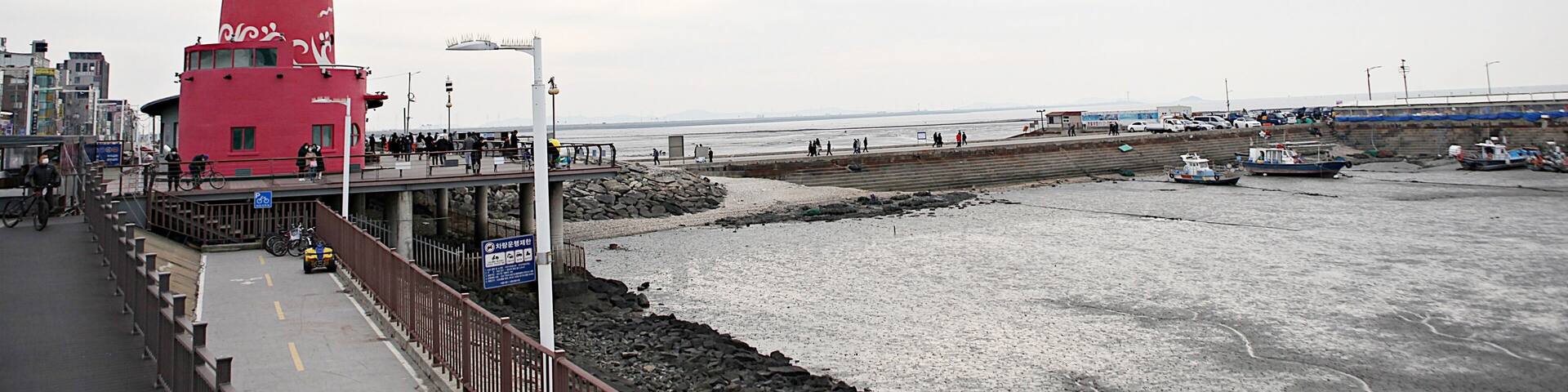 A red lighthouse, a rocky shoreline, and a pier with people and boats.