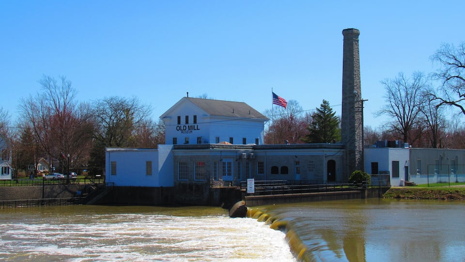 The Old Mill Museum with a large chimney, a flowing river, and an American flag.