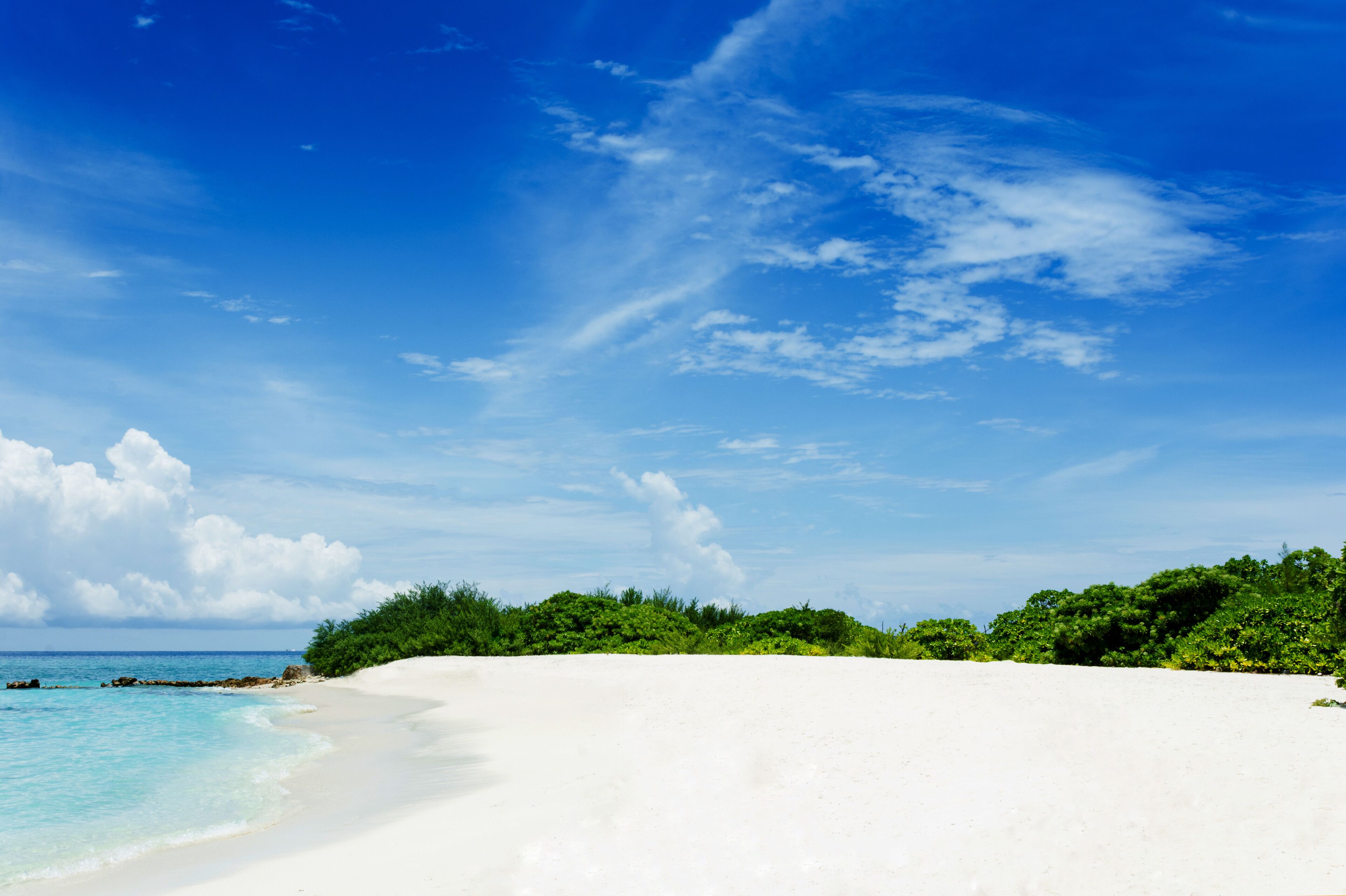 A white sandy beach with clear turquoise water and a lush green forest in the background under a blue sky with scattered clouds.