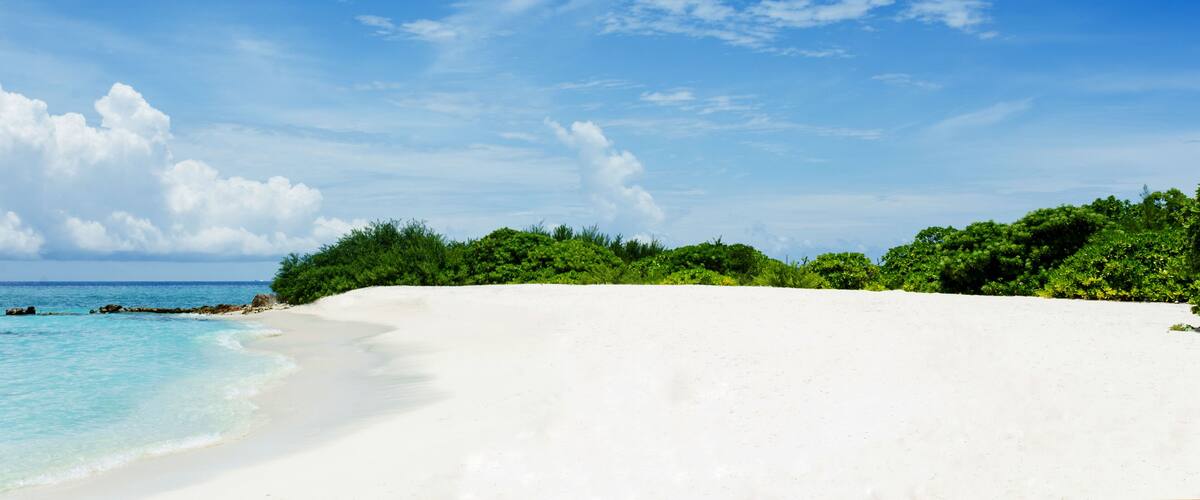 A white sandy beach with clear turquoise water and a lush green forest in the background under a blue sky with scattered clouds.