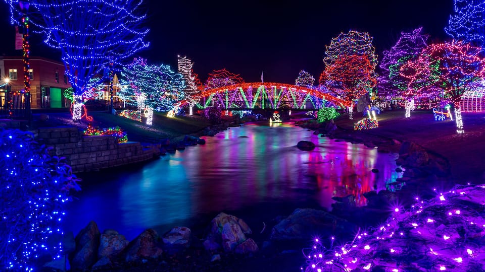 A river with a bridge adorned with colorful lights, surrounded by illuminated trees and a building with festive decorations.