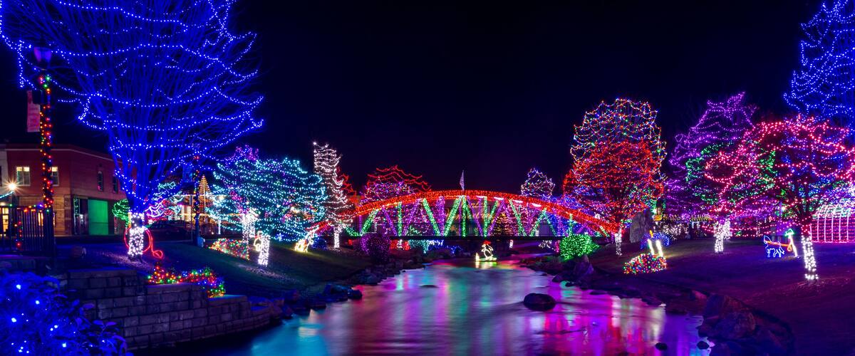 A river with a bridge adorned with colorful lights, surrounded by illuminated trees and a building with festive decorations.