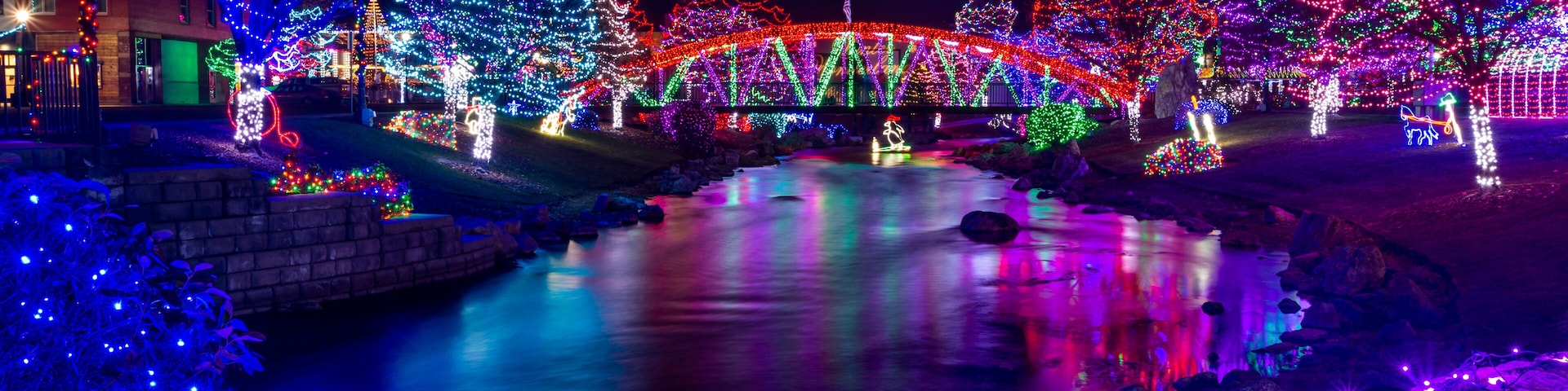 A river with a bridge adorned with colorful lights, surrounded by illuminated trees and a building with festive decorations.