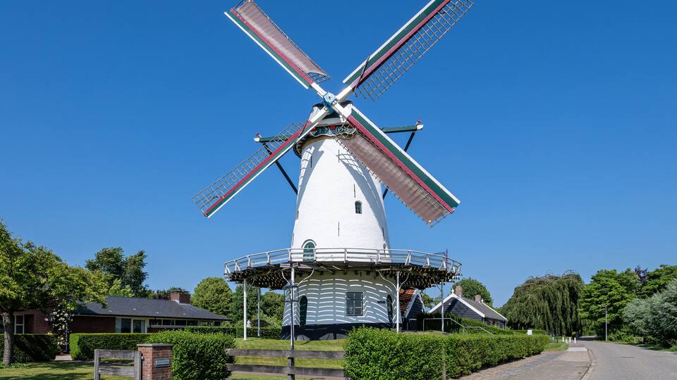 A white windmill with red and green stripes, surrounded by greenery and a clear blue sky.