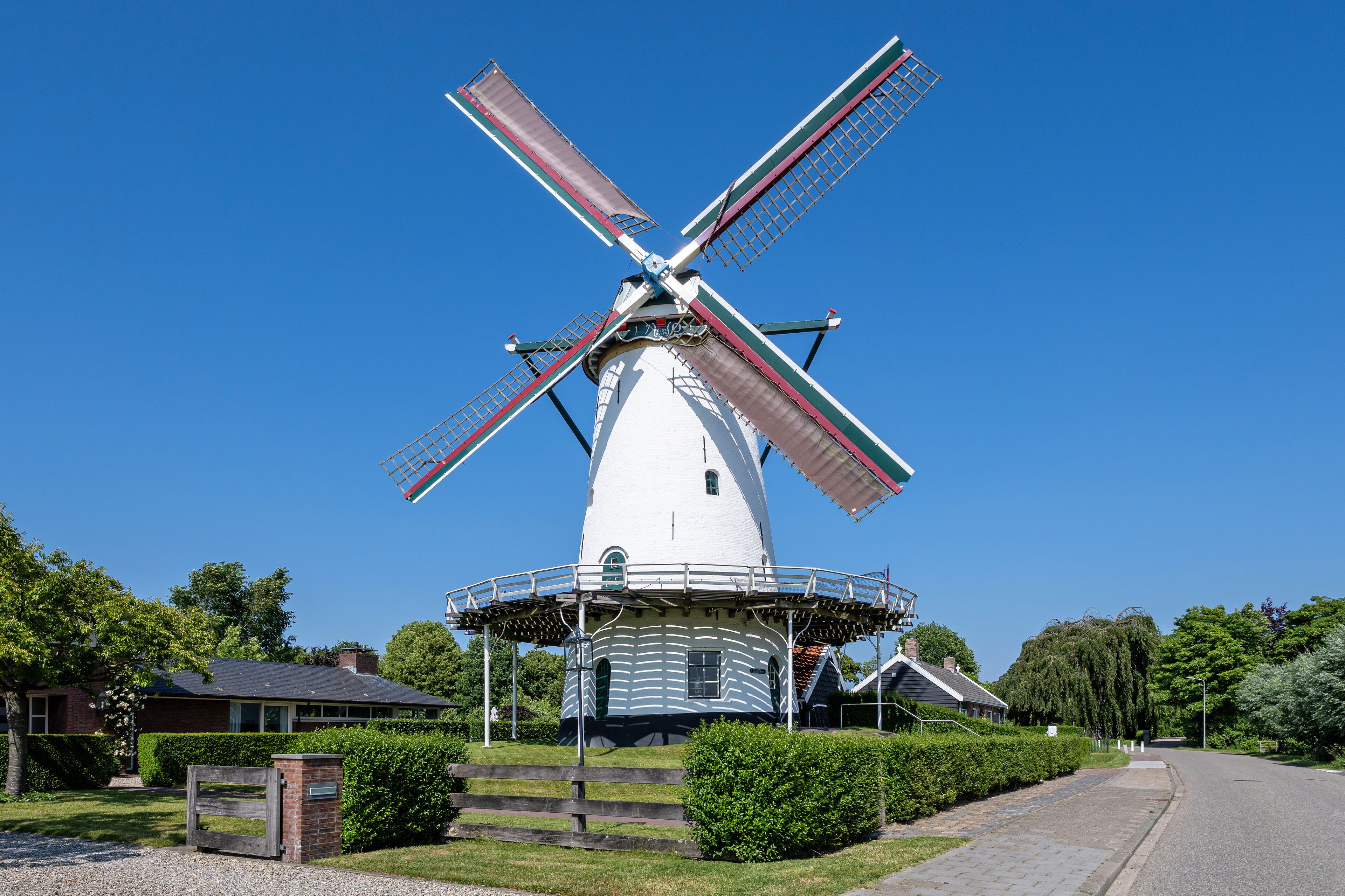 A white windmill with red and green stripes, surrounded by greenery and a clear blue sky.