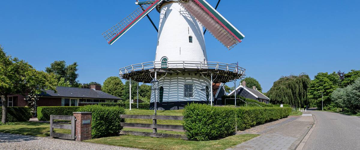 A white windmill with red and green stripes, surrounded by greenery and a clear blue sky.