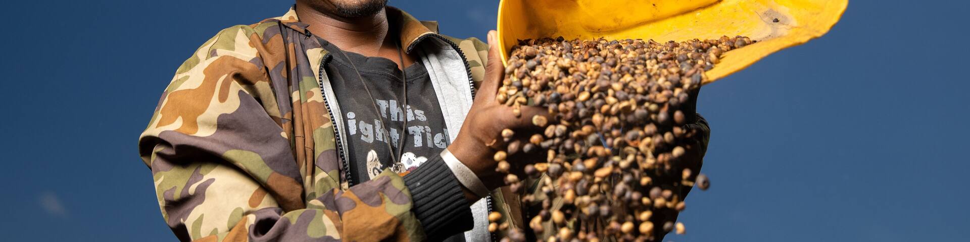 A person holding a yellow container filled with coffee beans against a clear blue sky.