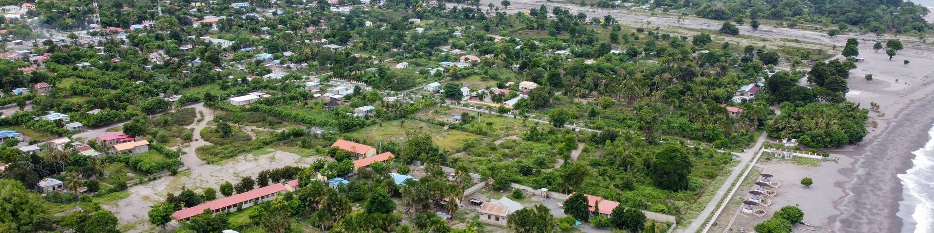 Panoramic view of green, tree covered landscape, houses, buildings, roads and black sandy beach, aerial drone views of Liquica town in Timor Leste, Southeast Asia