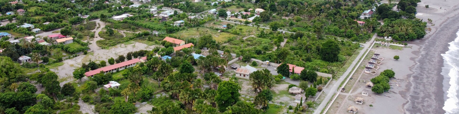 Panoramic view of green, tree covered landscape, houses, buildings, roads and black sandy beach, aerial drone views of Liquica town in Timor Leste, Southeast Asia