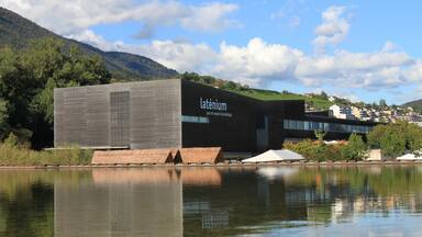 Un edificio moderno con el nombre 'Latenius' en su fachada, ubicado junto a una masa de agua tranquila con una montaña al fondo.
