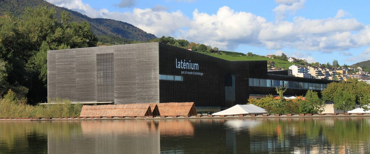 A modern building with the name 'Latenius' on its facade, situated by a calm body of water with a mountain in the background.