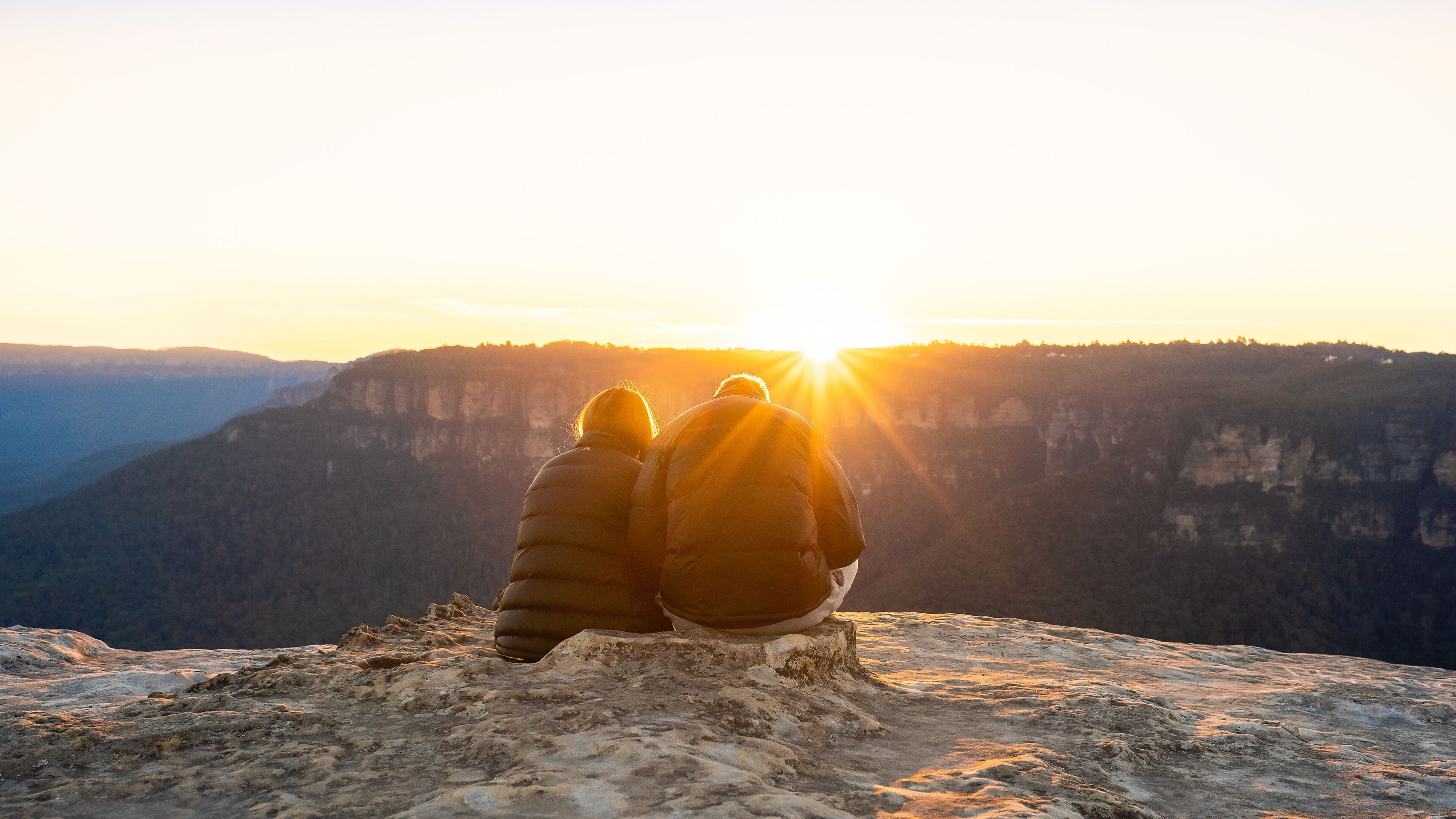 Duas pessoas sentadas em um afloramento rochoso com vista para um vale, com uma cordilheira ao fundo e o sol se pondo.