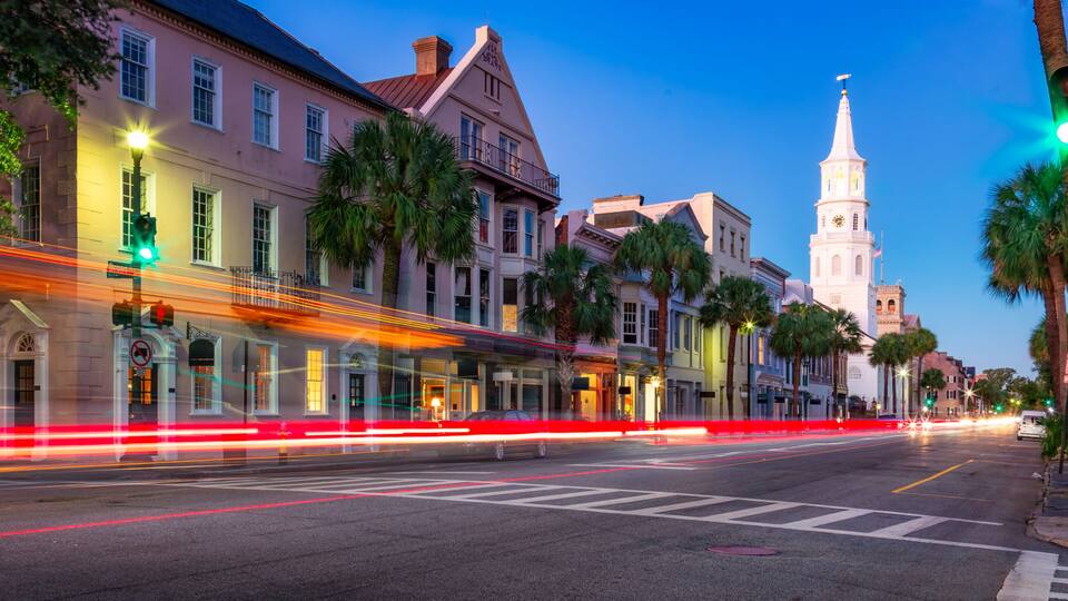 A city street at dusk with historic buildings, a church tower, and light trails from vehicles.