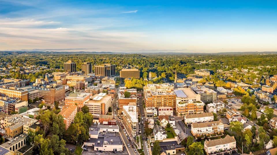 An aerial view of a city with a mix of modern and historic buildings, surrounded by green spaces and a distant mountain range.