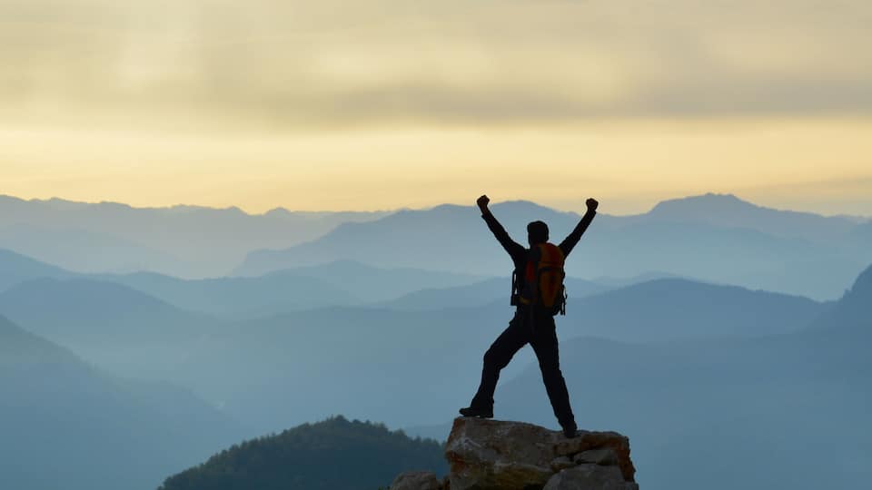 A person standing on a rock with arms raised, surrounded by mountain ranges.