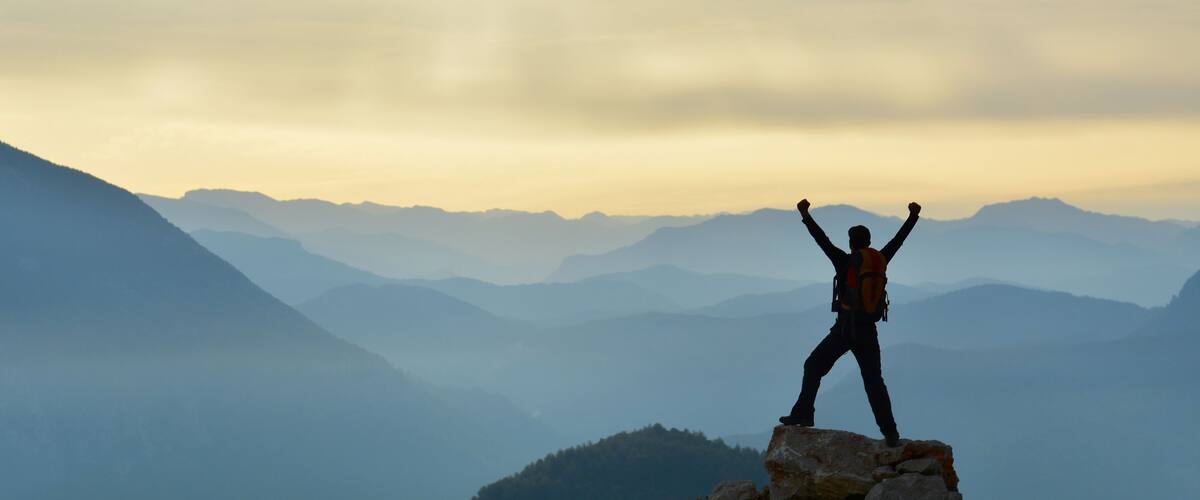 A person standing on a rock with arms raised, surrounded by mountain ranges.