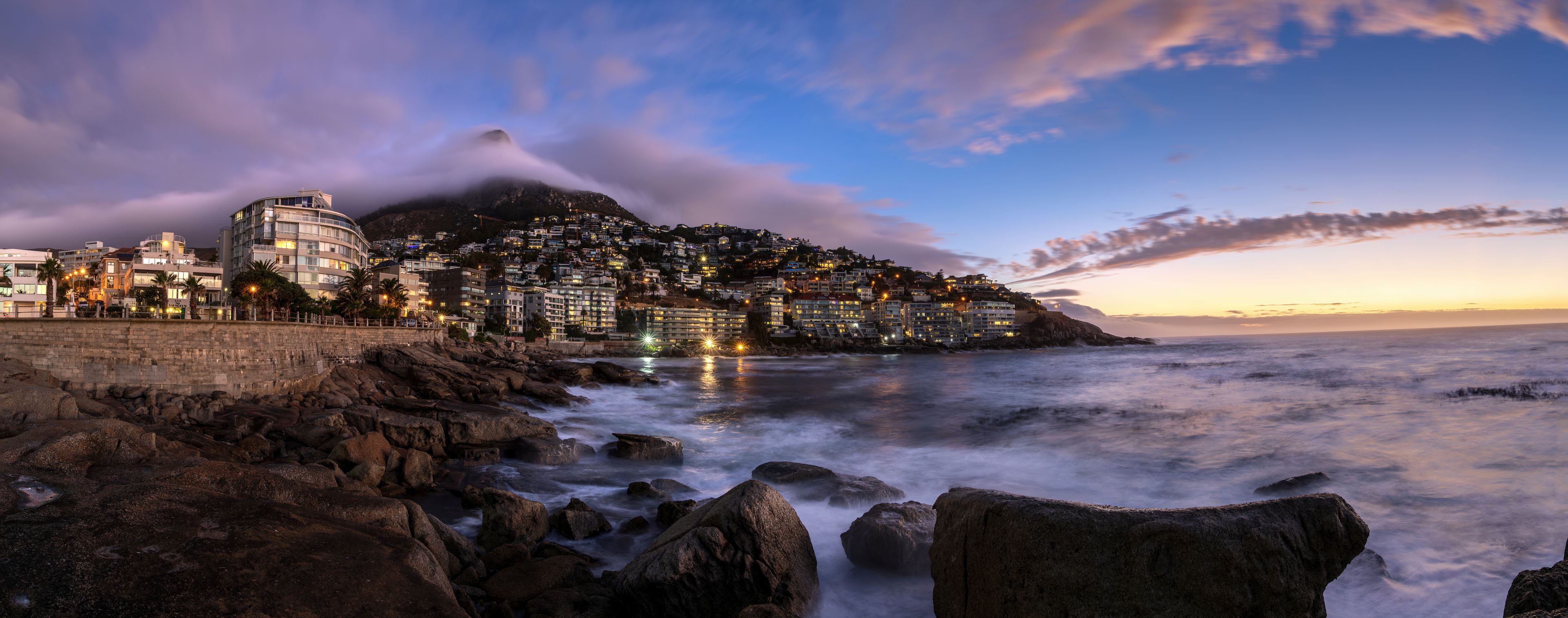 Panoramic view of Bantry Bay city lights at sunset on Sea Point Promenade with dramatic low cloud rolling over Lion’s Head mountain in background, Saunder's Rock beach, Cape Town, South Africa.
