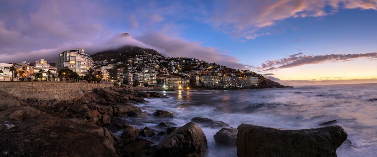 A coastal cityscape at dusk with a mountain in the background and a rocky shoreline in the foreground.