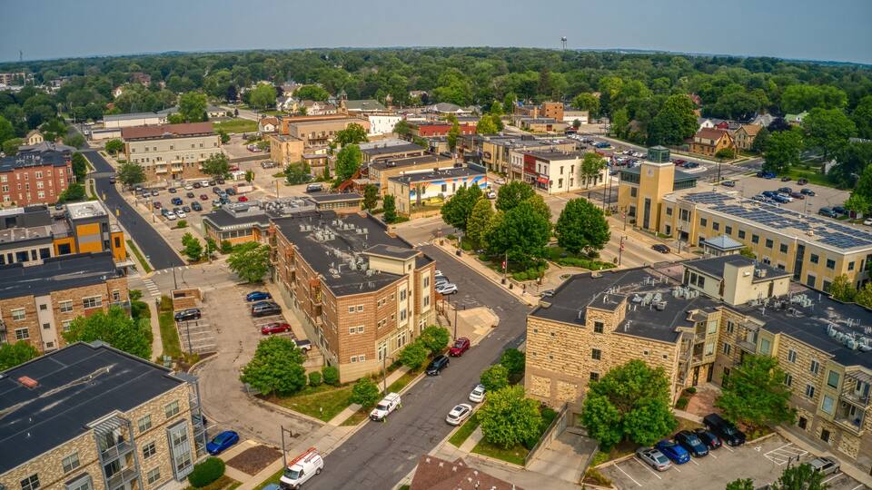 An aerial view of a town with a mix of residential and commercial buildings, a main road, and surrounding greenery.
