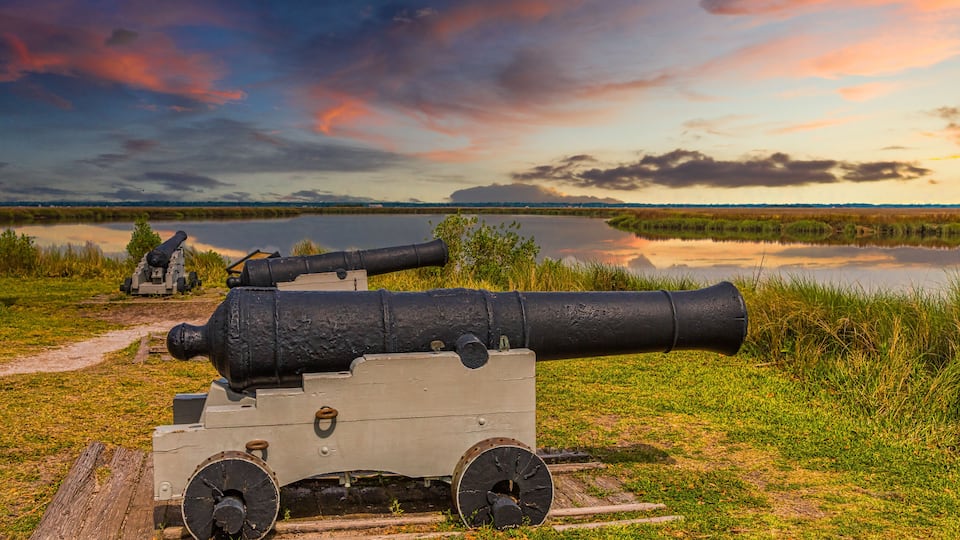A cannon on a grassy field with a lake and mountains in the background.