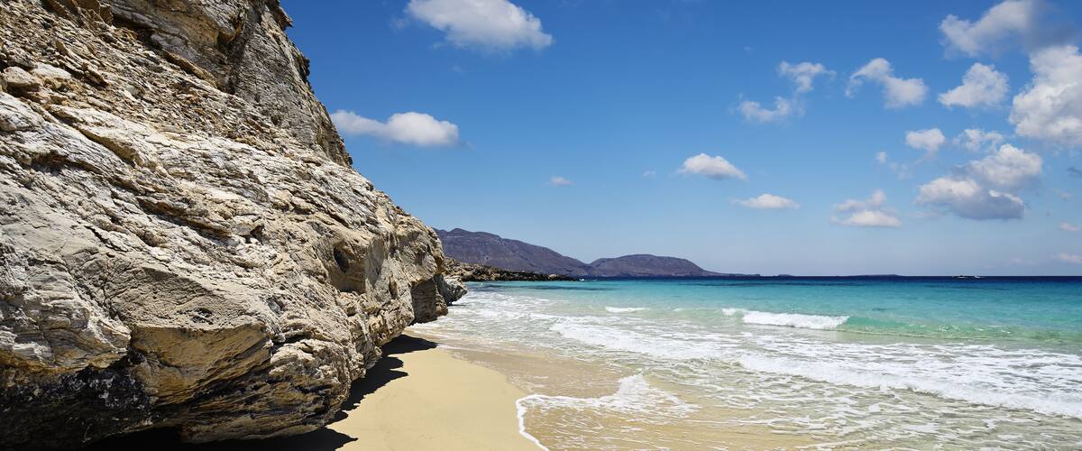A sandy beach with clear turquoise water, a rocky cliff, and distant mountains under a blue sky.