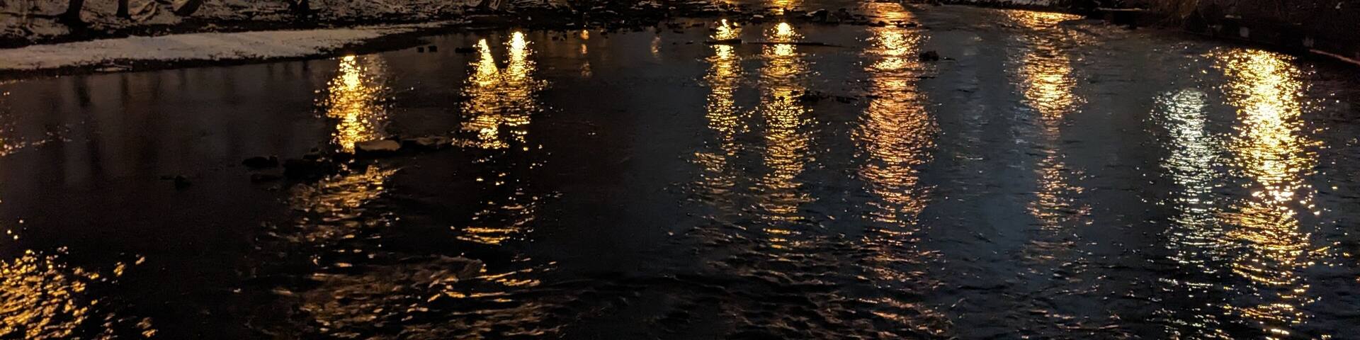 Un río que refleja luces, rodeado de árboles desnudos y un sendero cubierto de nieve.