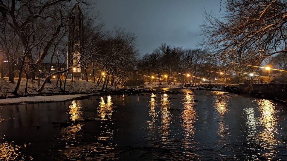 A river reflecting lights, surrounded by bare trees and a snow-covered path.