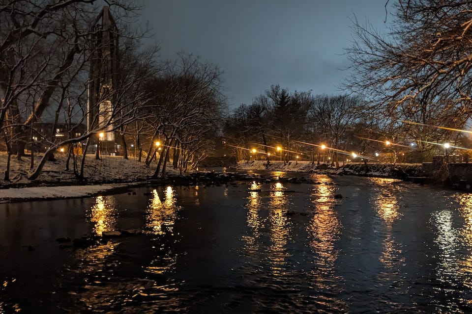 Een rivier die lichtjes weerspiegelt, omringd door kale bomen en een met sneeuw bedekte pad.