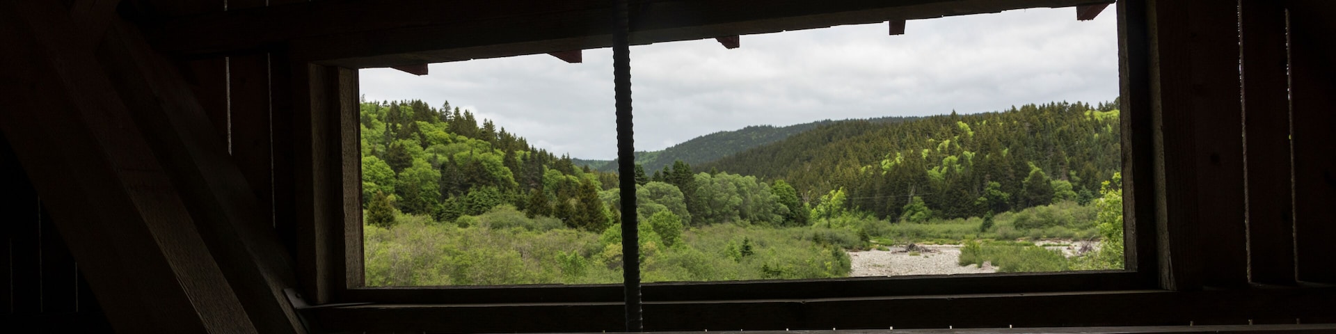 A view through a wooden window of a forested valley with a river and distant hills.