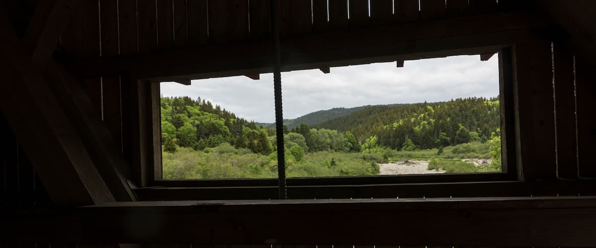 A view through a wooden window of a forested valley with a river and distant hills.