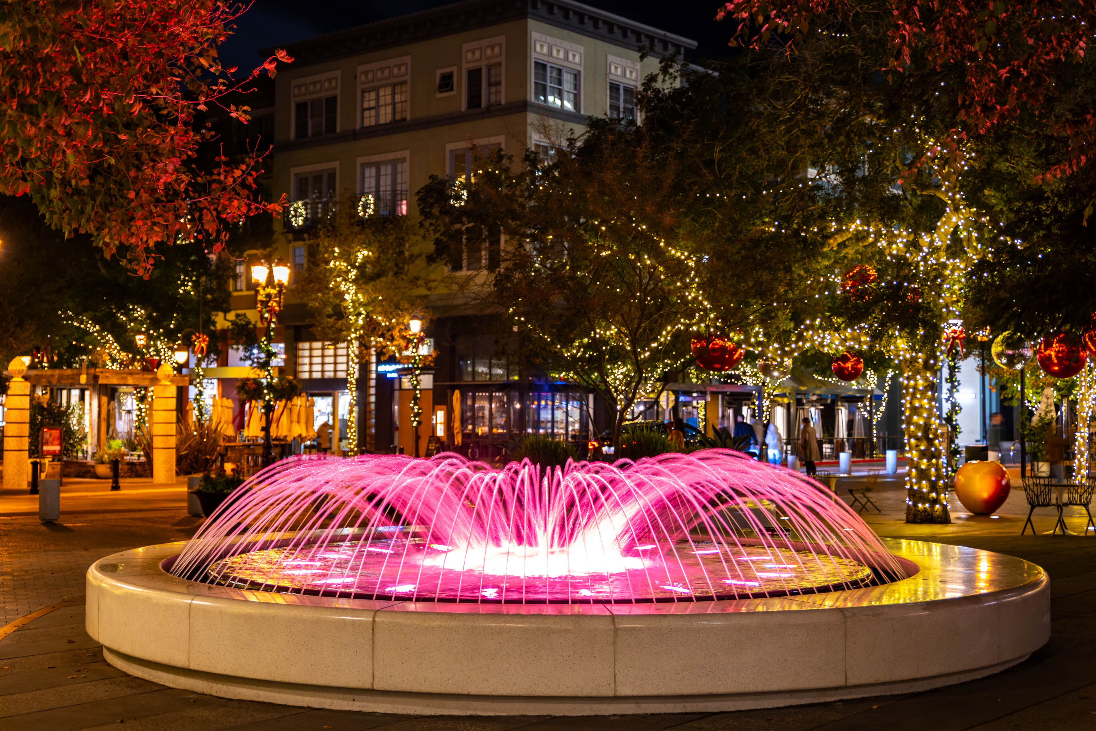 A fountain with water jets, surrounded by trees and buildings with illuminated windows.