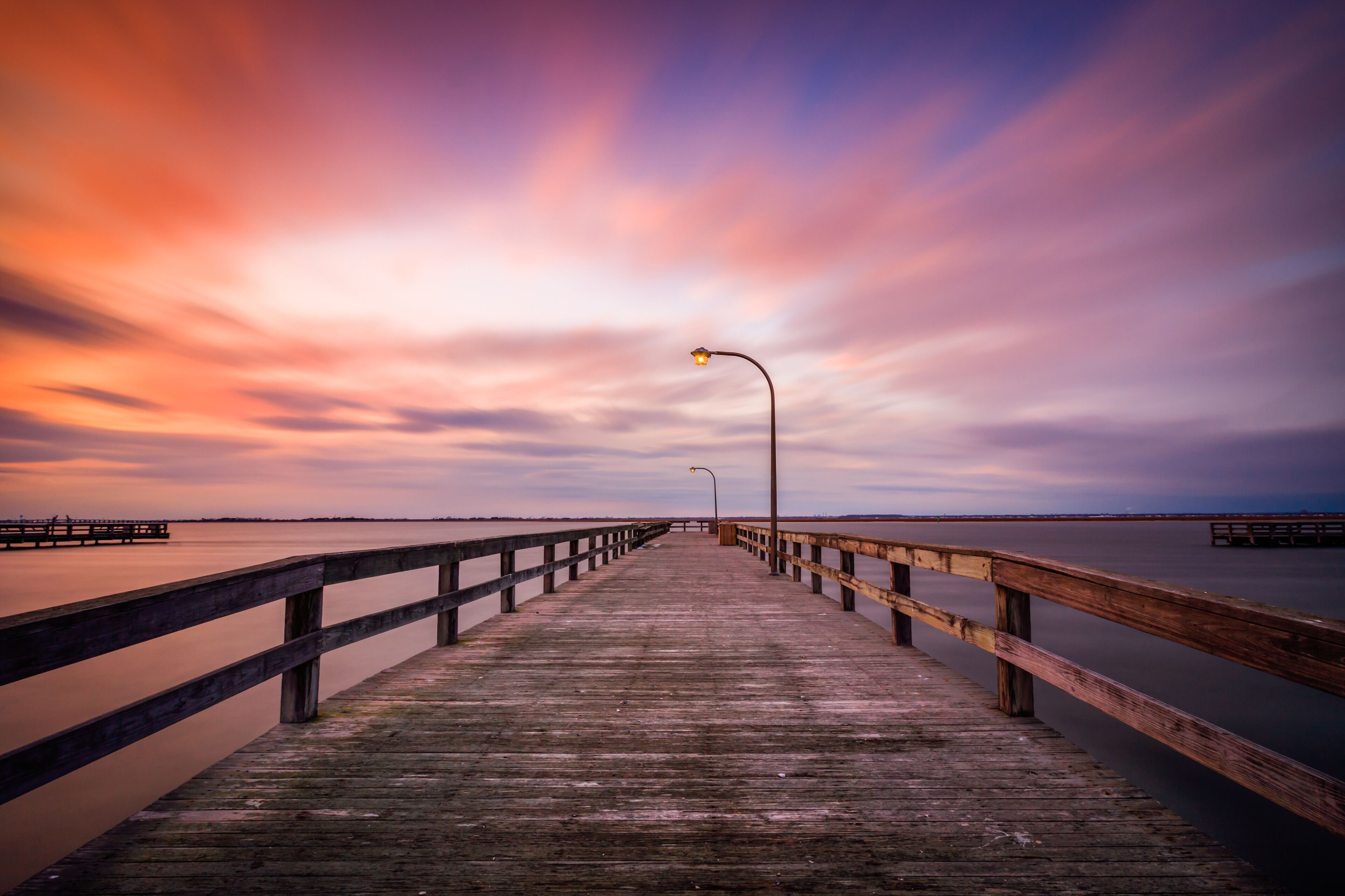 Un muelle de madera que se adentra en aguas tranquilas, con un cielo al atardecer y una costa lejana.