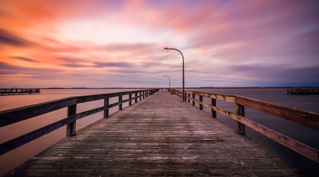 A wooden pier extending into calm waters, with a sunset sky and a distant shoreline.