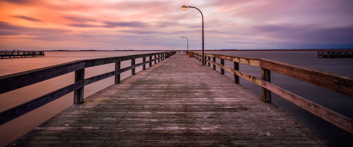 Une jetée en bois s’étendant sur des eaux calmes, avec un ciel au coucher du soleil et une côte lointaine.