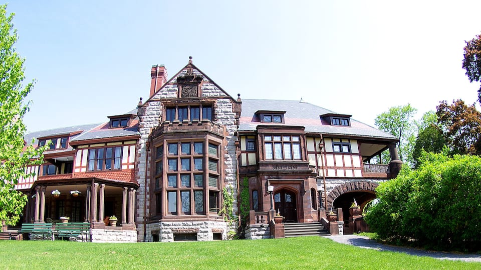 A large, historic house with a prominent tower, surrounded by lush greenery and a well-maintained lawn.