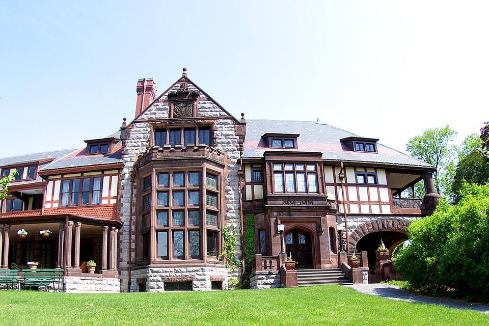 Uma casa grande e histórica com uma torre imponente, rodeada por vegetação exuberante e um gramado bem cuidado.