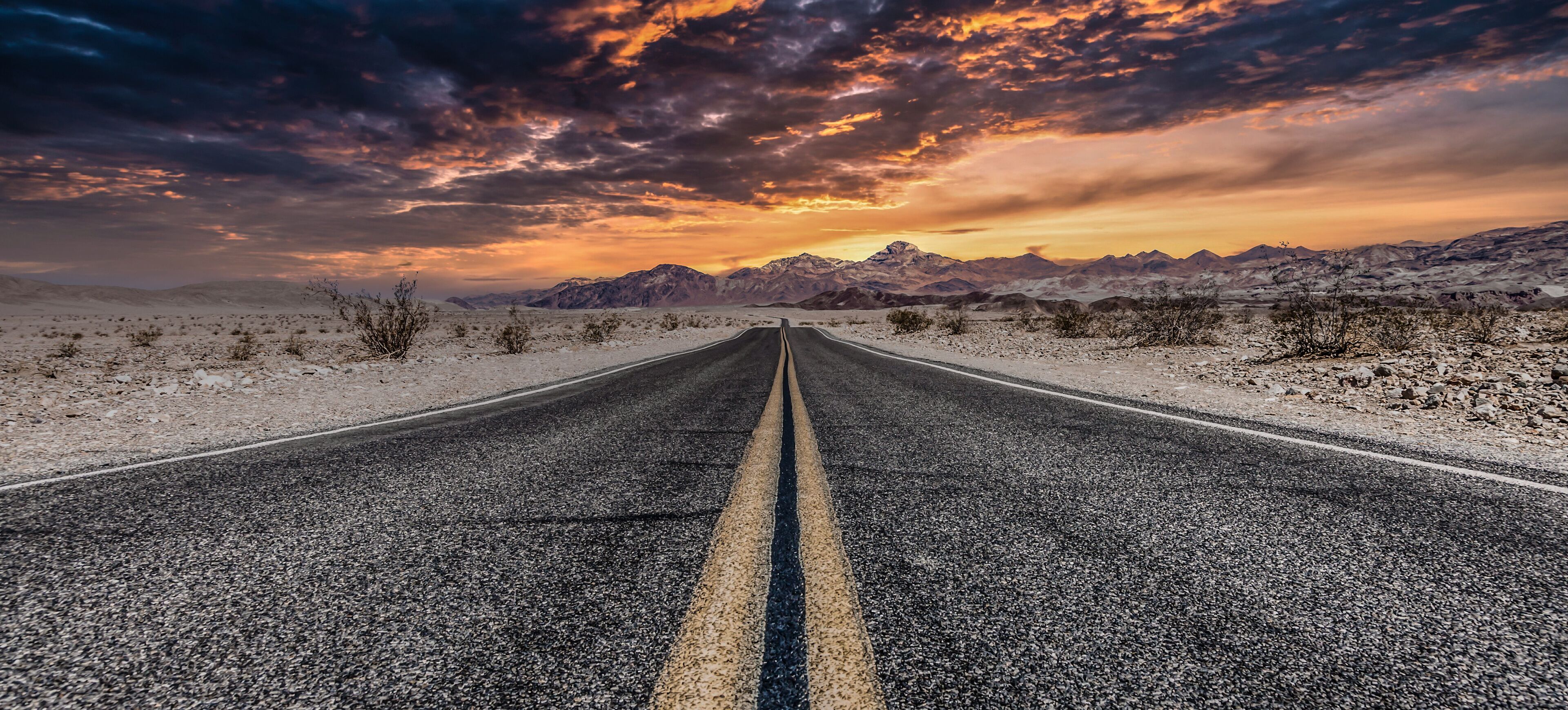 A road in a desert landscape with mountains in the distance and a dramatic sunset sky.