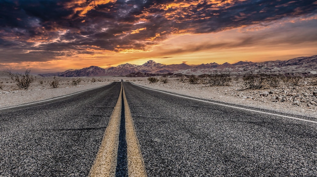 A road in a desert landscape with mountains in the distance and a dramatic sunset sky.