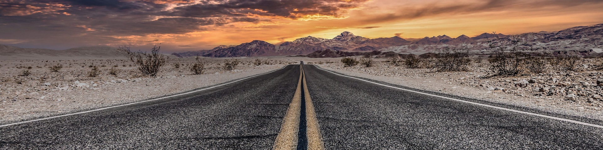 A road in a desert landscape with mountains in the distance and a dramatic sunset sky.
