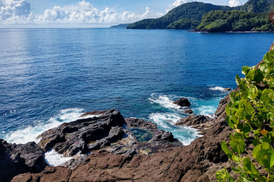 A coastal landscape with rocky shores, clear blue sea, and green hills in the background.